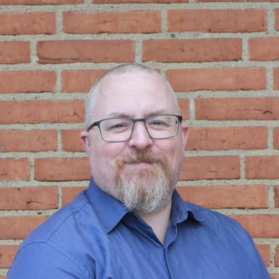 Alex Holst (he/him) Close-up photo of old white man in a blue shirt and with greying beard, very little hair and glasses standing in front of a red brick wall.